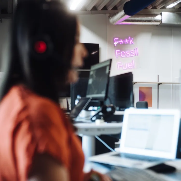 Frau mit Kopfhörern am Laptop im Büro, Neon-Schrift „F* Fossil Fuel“ im Hintergrund – Symbol für Energiewende